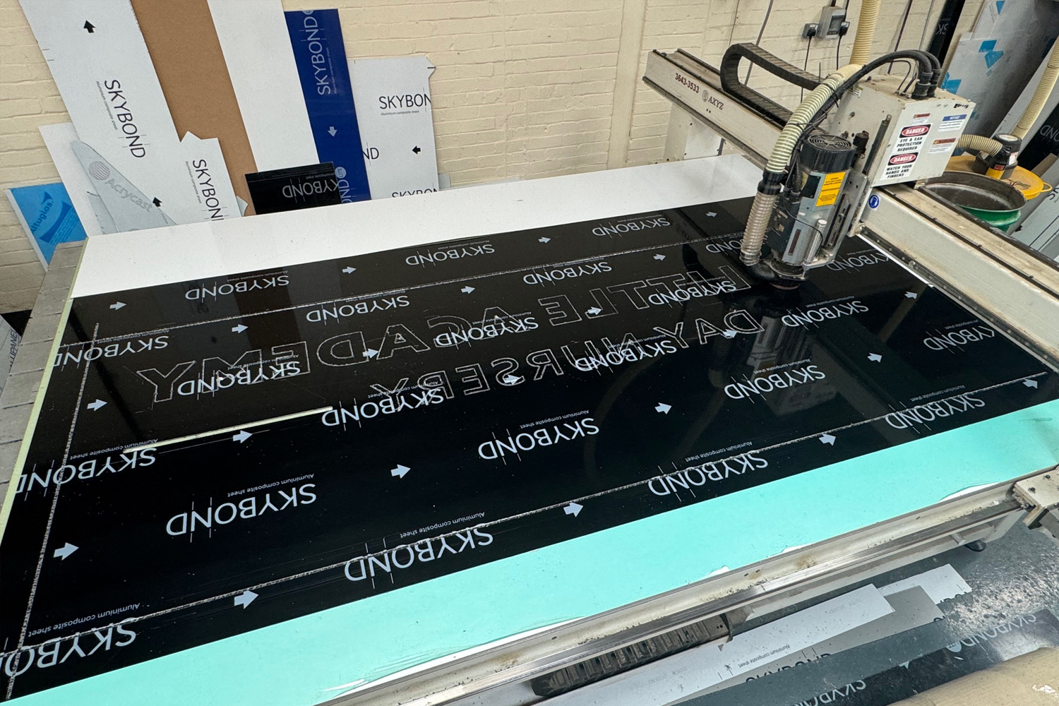 A wide shot showing a CNC router engraving letters and outlines into a black Skybond aluminium composite panel, used for custom exterior signage